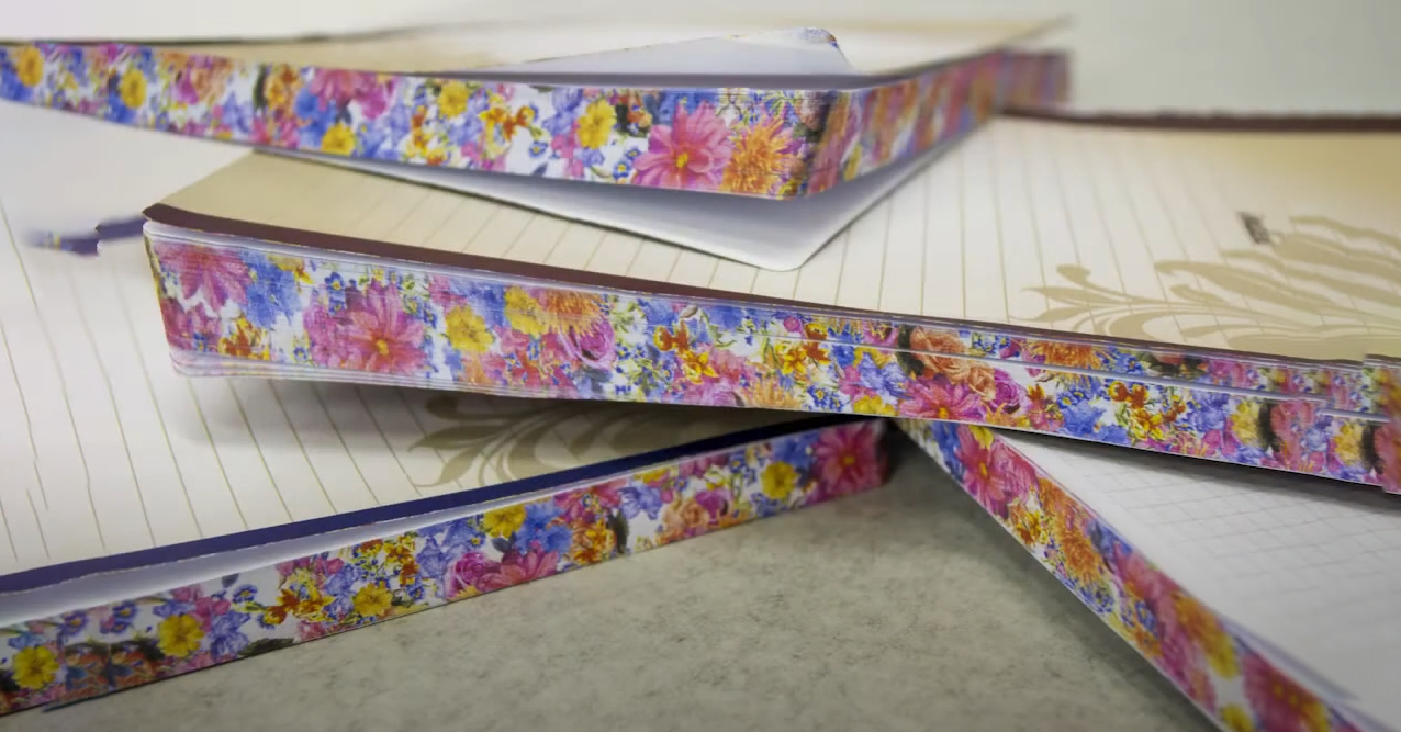 The image shows various notebooks stacked with floral border designs. The notebooks are open, revealing lined pages and decorative motifs with delicate patterns. The floral designs on the edges feature vibrant pink, yellow, and blue flowers, giving the notebooks a colorful and attractive appearance. The setting appears to be on a smooth, neutral-colored surface, possibly a desk. The overall image highlights aesthetically pleasing stationery with a focus on vivid floral decorations.